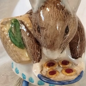 Peut inclure: Une figurine en céramique d'une souris brune vêtue d'une robe bleu et blanc et d'un chapeau de paille. La souris tient une assiette avec quatre biscuits rouges et jaunes.