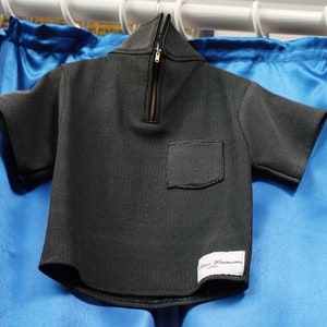 Peut inclure: Une chemise à manches courtes à motifs noirs et blancs avec un col zippé quart de tour et une petite poche. La chemise porte une étiquette avec le texte "Alan Woomam London". La chemise est suspendue à une tringle blanche.