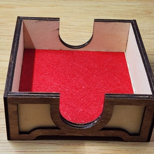 Puede incluir: Una bandeja de dados de madera con un forro de fieltro rojo. La bandeja tiene una secci&oacute;n elevada en el centro para evitar que los dados se salgan.