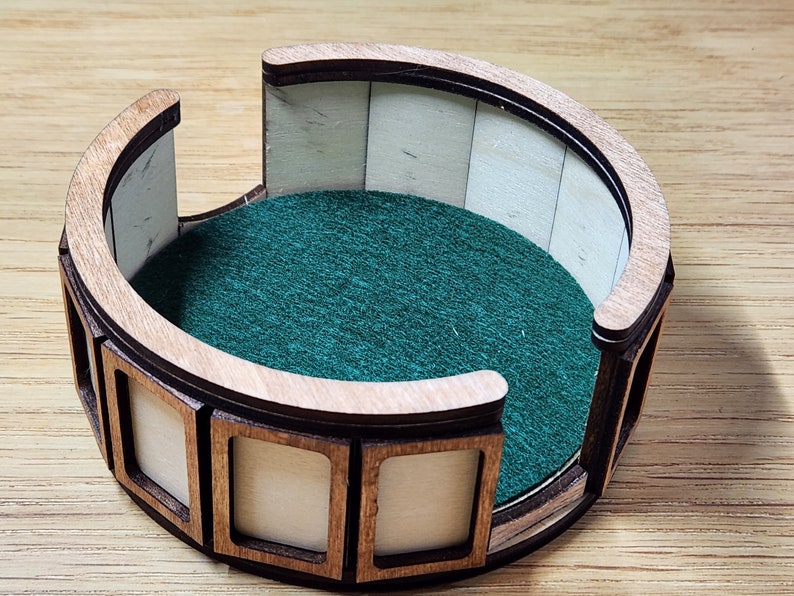 Pu&ograve; includere: Un vassoio per dadi in legno con un rivestimento in feltro verde e un bordo decorativo. Il vassoio ha otto scomparti attorno al bordo.