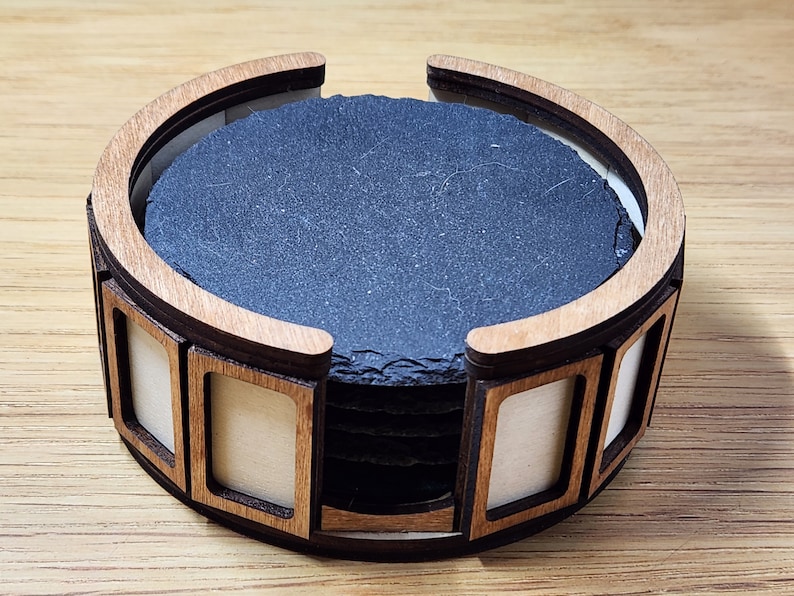 Pu&ograve; includere: Un set di sei sottobicchieri in ardesia nera in un portaoggetti in legno con finitura in legno naturale. Il portaoggetti ha un design curvo e presenta un ritaglio su ogni lato.