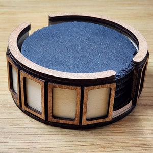 Pu&ograve; includere: Un set di sei sottobicchieri in ardesia nera in un supporto in legno con una cornice decorativa. Il supporto ha una forma arrotondata ed &egrave; in legno marrone chiaro.
