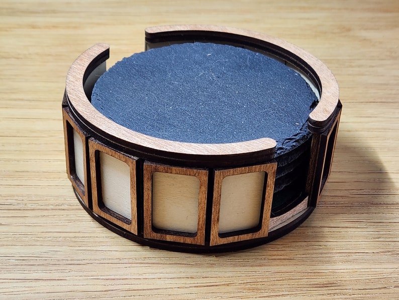 Pu&ograve; includere: Un set di sei sottobicchieri in ardesia nera in un supporto in legno con una cornice decorativa. Il supporto ha una forma arrotondata ed &egrave; in legno marrone chiaro.