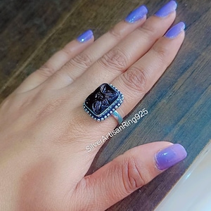 May include: A silver ring with a rectangular, black stone featuring a floral design. The stone is set in a silver frame with a beaded edge. The ring is on a hand with purple nail polish. The text "SilverArtisanRing925" is visible.