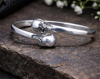 May include: A sterling silver bracelet with skull accents. The bracelet features two skull designs at each end of the open cuff. The jewelry is displayed on a dark, textured surface. The text on the image reads "925 Sterling Silver Skull Bracelet Handmade Gothic Jewelry."