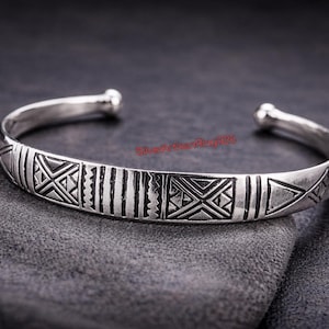 May include: Silver cuff bracelet with geometric patterns. The open-ended bracelet features etched designs, including triangles, lines, and cross-like shapes. The bracelet is displayed on a dark grey surface.
