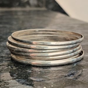 Op de afbeelding: Een stapel van zeven zilveren armbanden, elk met een iets andere breedte en een gladde, gepolijste afwerking.