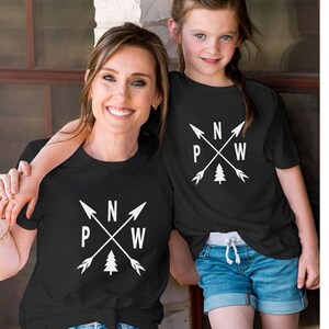 May include: A black t-shirt with a white compass design featuring arrows pointing north, west, east, and south. The design also includes a small pine tree. The t-shirt is being worn by two people, an adult and a child.
