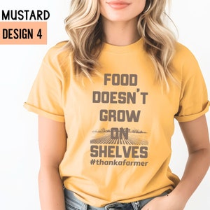 May include: A mustard yellow t-shirt with the text "Food Doesn't Grow On Shelves #thankafarmers" in brown text. The shirt has a field of crops graphic with a brown border.