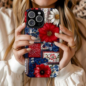 May include: A floral phone case featuring a patchwork design with red, white, and blue floral patterns. The case has a red gerbera daisy and a white daisy. The phone case is held by a person.