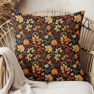 May include: A square throw pillow featuring a dark background with a repeating pattern of autumn leaves in shades of yellow, orange, and red, interspersed with small blue berries and dark green foliage, creating a dense botanical design.