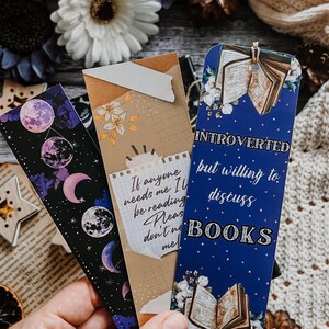 May include: Three decorative bookmarks with unique designs. One features a celestial moon phase pattern, another has a quote, and the third reads "Introverted but willing to discuss BOOKS." Each bookmark is rectangular and colorful.