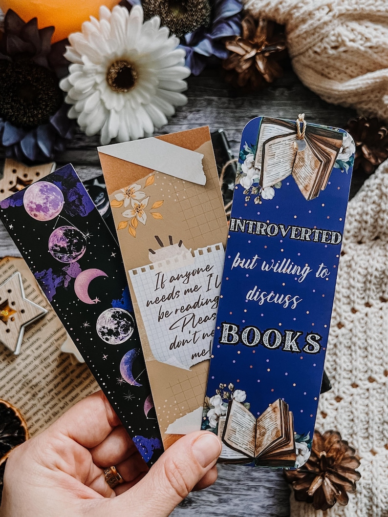 May include: Three decorative bookmarks with unique designs. One features a celestial moon phase pattern, another has a quote, and the third reads "Introverted but willing to discuss BOOKS." Each bookmark is rectangular and colorful.