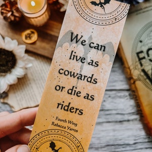 May include: A bookmark with a dragon design and the quote "We can live as cowards or die as riders." from the book "Fourth Wing" by Rebecca Yarros.