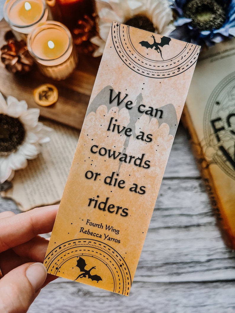 May include: A bookmark with a dragon design and the quote "We can live as cowards or die as riders." from the book "Fourth Wing" by Rebecca Yarros.