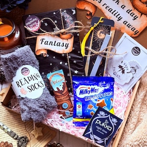 May include: A gift box containing a pair of gray fuzzy reading socks, a black wrapped book labeled Fantasy, a bookmark with a yellow tassel, a Milky Way Magic Stars candy bag, a Pukka Night Time tea bag, and a hot chocolate sachet, with a sign reading A book a day keeps reality away.