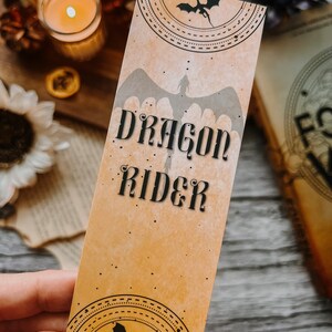 May include: A tan bookmark with a dragon silhouette and the text "Dragon Rider" in black lettering. The bookmark has a decorative border with a smaller dragon silhouette at the bottom.