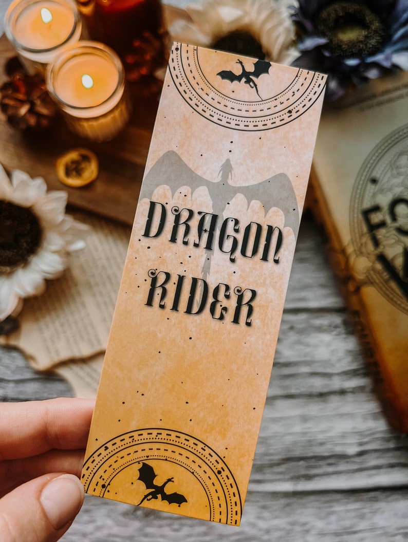 May include: A tan bookmark with a dragon silhouette and the text "Dragon Rider" in black lettering. The bookmark has a decorative border with a smaller dragon silhouette at the bottom.