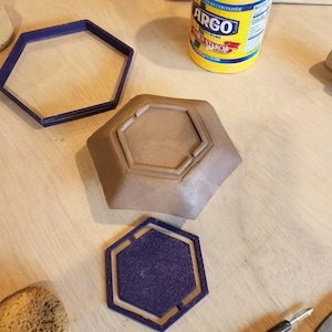 Może przedstawiać: Brązowa, sześciokątna ceramiczna miska z pasującym fioletowym, sześciokątnym podkładem i fioletową, sześciokątną formą. Żółty pojemnik ze skrobią kukurydzianą Argo znajduje się w tle. Przedmioty znajdują się na jasnej drewnianej powierzchni.