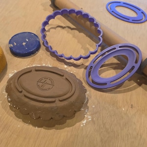 Könnte beinhalten: Eine Sammlung von Backutensilien auf einer Holzoberfläche. Enthält einen braunen, ovalen Ausstecher mit gewelltem Rand, einen lila Ausstecher, ein Nudelholz und einen blauen Deckel. Der Ausstecher hat den Text "GOOD IRON KITCHEN" und "KITCHENWARE CA".