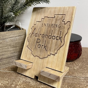 May include: A wooden sign with the words "ENTERING Adirondack Park" carved into the surface. The sign is light brown and features a map outline. It is propped up by two wooden supports.