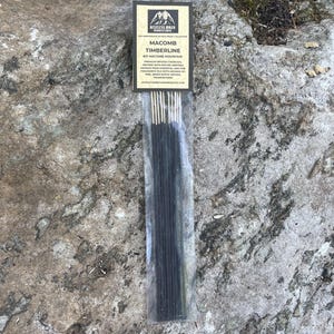 May include: A package of incense sticks on a rock. The label reads "Macomb Timberline #21 Macomb Mountain" and describes the incense as premium infused charcoal aromas from essential and fine fragrance oils with aromas of pine, warm rustic woods, and frankincense.