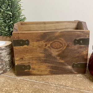 May include: A rectangular wooden storage box with a dark brown stained finish and visible wood grain. The front panel features four decorative metal corner plates in a muted bronze tone, each secured with two small black screws at the corners.