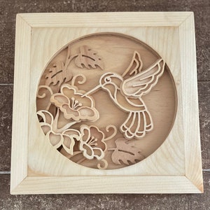May include: A wooden wall art piece with a cutout design of a hummingbird with its beak in a flower. The hummingbird and flower are surrounded by leaves and vines.