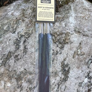 May include: A package of incense sticks labeled "MIST & GRANITE #7 GRAY PEAK" with a mountain logo. The incense sticks are dark gray and are in a clear plastic sleeve. The background is a gray rock.