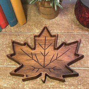 May include: A wooden maple leaf-shaped tray with a carved leaf vein design. The tray is a warm brown color and has a raised edge. The tray is likely used for decorative purposes or serving small items.