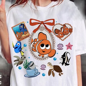 Könnte beinhalten: Weißes T-Shirt mit einem farbenfrohen Aufdruck von Figuren aus dem Film "Findet Nemo". Das Design zeigt Nemo, Dorie und andere Meeresbewohner in einer verspielten Anordnung. Das Shirt hat einen klassischen Rundhalsausschnitt und kurze Ärmel.