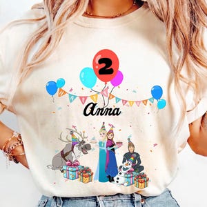 May include: Ivory t-shirt with a birthday graphic. The design includes balloons, the number 2, and the name "Anna." Cartoon characters and gift boxes are also featured, making it suitable for a child's second birthday celebration.