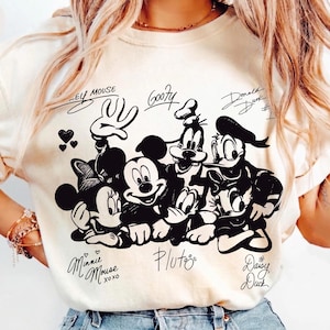 May include: Ivory-colored t-shirt featuring a black and white graphic of Mickey Mouse, Minnie Mouse, Goofy, Donald Duck, and Pluto. The shirt includes the characters' signatures and the text "Colors Ivory".