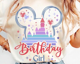 May include: Cream-colored t-shirt with a colorful graphic design. The design features a castle, Mickey Mouse ears, balloons, stars, and the words "Birthday Girl" in a script font. The overall theme is celebratory and whimsical.