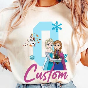 May include: Ivory-colored t-shirt featuring a custom design with the word "Custom" in a cursive font. The design includes the number "0" in light blue, snowflakes, and illustrations of Elsa, Anna, and Olaf from the movie Frozen.