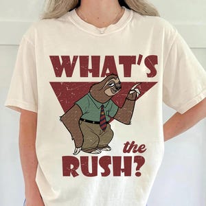May include: Cream-colored t-shirt featuring a cartoon sloth in a green shirt, brown pants, and a red tie. The text "WHAT'S the RUSH?" is printed in a distressed red font, with the word "WHAT'S" above the sloth and "the RUSH?" below.