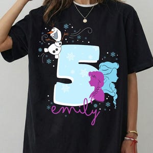 Puede incluir: Camiseta negra con un dise&ntilde;o gr&aacute;fico que presenta el n&uacute;mero 5 en azul claro, copos de nieve y siluetas de personajes. El nombre "emily" est&aacute; escrito en cursiva. Tambi&eacute;n se incluye un mu&ntilde;eco de nieve de dibujos animados.