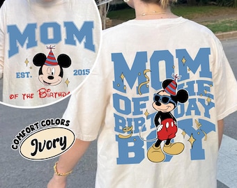 May include: Ivory-colored t-shirt with a blue "MOM" graphic and a Mickey Mouse illustration wearing sunglasses and a party hat. The shirt also features the text "EST. 2015" and "OF THE BIRTHDAY BOY". The shirt is part of the Comfort Colors collection.