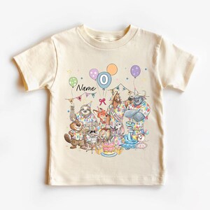 May include: Cream-colored t-shirt featuring a colorful birthday party scene with cartoon animals. The design includes balloons, a cake, and the number 0. The word "Name" is also visible.