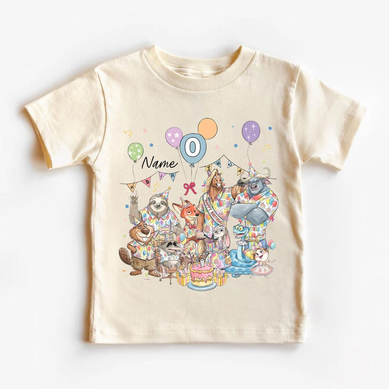 May include: Cream-colored t-shirt featuring a colorful birthday party scene with cartoon animals. The design includes balloons, a cake, and the number 0. The word "Name" is also visible.