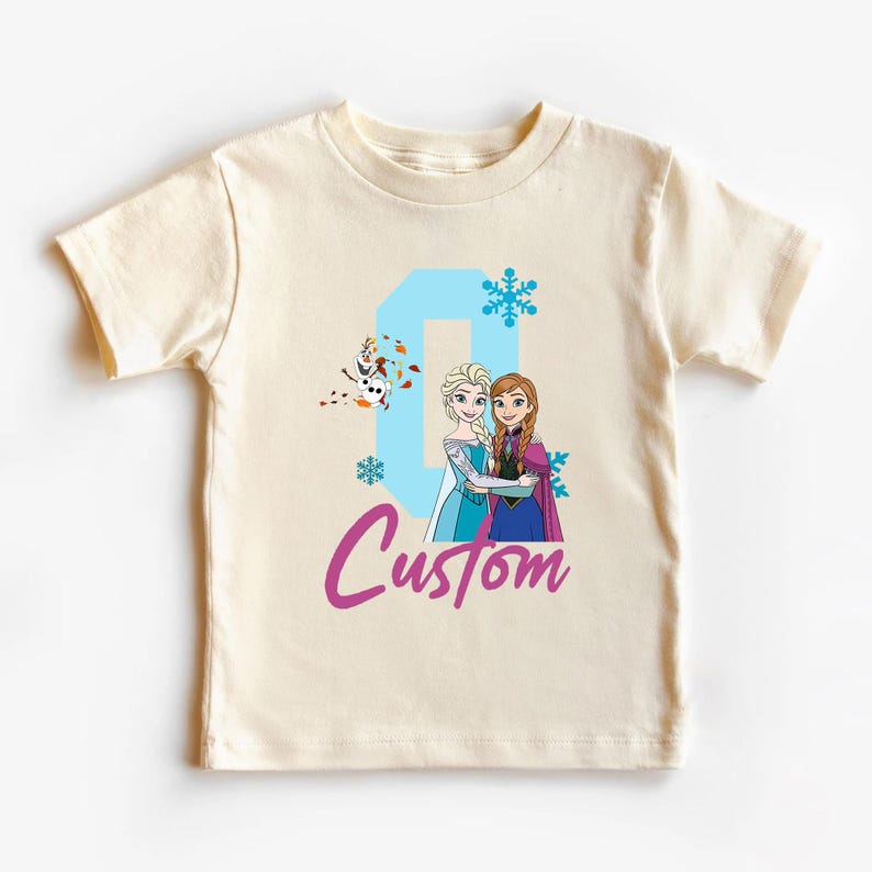May include: Cream-colored t-shirt featuring a graphic of two animated princesses, a snowman, snowflakes, and the word "Custom" in a script font. The number "0" is in light blue.