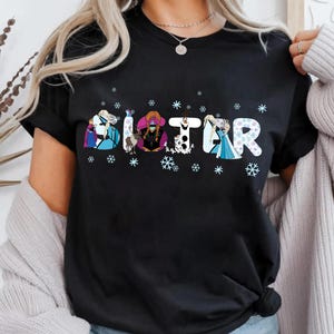 May include: Black t-shirt with the word "SISTER" in a whimsical font, each letter featuring a different cartoon character. Snowflakes are scattered around the letters. The shirt is a casual wear item.