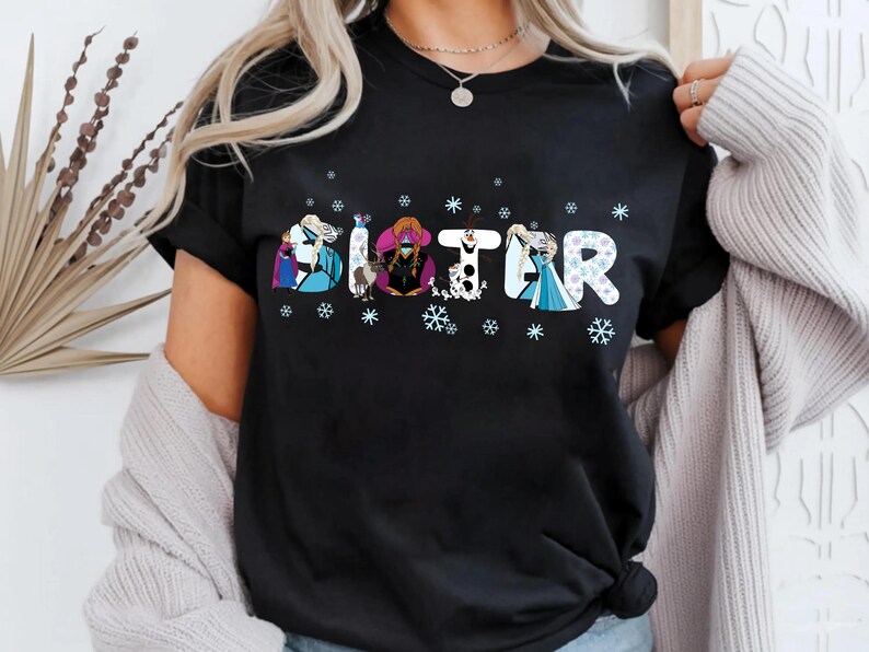 May include: Black t-shirt with the word "SISTER" in a whimsical font, each letter featuring a different cartoon character. Snowflakes are scattered around the letters. The shirt is a casual wear item.
