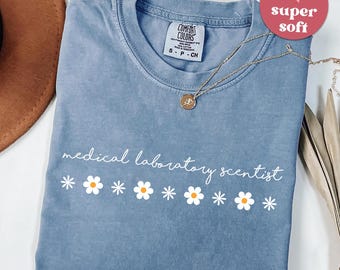 May include: A blue jean colored t-shirt with the words "medical laboratory scientist" in white script, along with a row of daisy graphics. A gold necklace with a pendant is draped across the shirt. A pink heart-shaped sticker says "super soft".