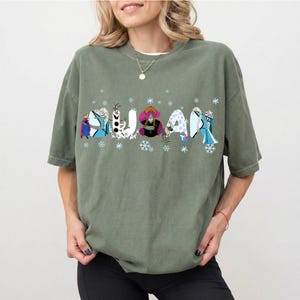 May include: Olive green t-shirt with the word "FROZEN" in a colorful, cartoon-style font. Each letter is decorated with characters and snowflakes from the animated movie. The shirt has short sleeves and a relaxed fit.