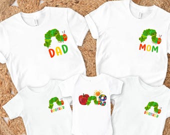 May include: White t-shirts and a onesie featuring a colorful caterpillar design. The shirts have the words "DAD", "MOM", and "BROTHER" printed on them. The onesie has the word "ONE" with a sun and apple graphic.