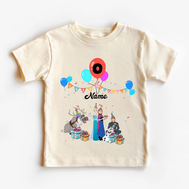 May include: Cream-coloured t-shirt with a colourful birthday design. The design includes balloons, a banner, and cartoon characters. The number "0" is displayed on a red balloon, and the word "Name" is written in cursive.