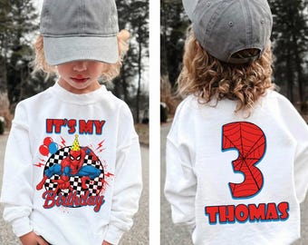 May include: White sweatshirt with a Spider-Man graphic, the text "It's My Birthday", and a checkered background. The back of the shirt features a red number 3 with a spiderweb design and the name "Thomas". The child is wearing a gray baseball cap.