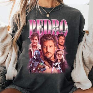 May include: Dark gray sweatshirt featuring a collage of images of Pedro Pascal, with the name "PEDRO Pascal" in a pink gradient font. The images include Pascal in various roles, including The Mandalorian and with Grogu.