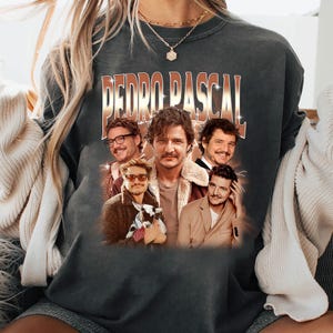Peut inclure: Sweat-shirt gris foncé avec un graphisme de Pedro Pascal et le texte "PEDRO PASCAL" dans un style vintage. Le graphisme comprend plusieurs images de l'acteur. Le sweat-shirt est de style col rond.